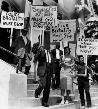 Protesters descending courthouse steps carrying signs reading 'Segregated Schools Must Go Now' and 'Police Brutality Must Stop'
